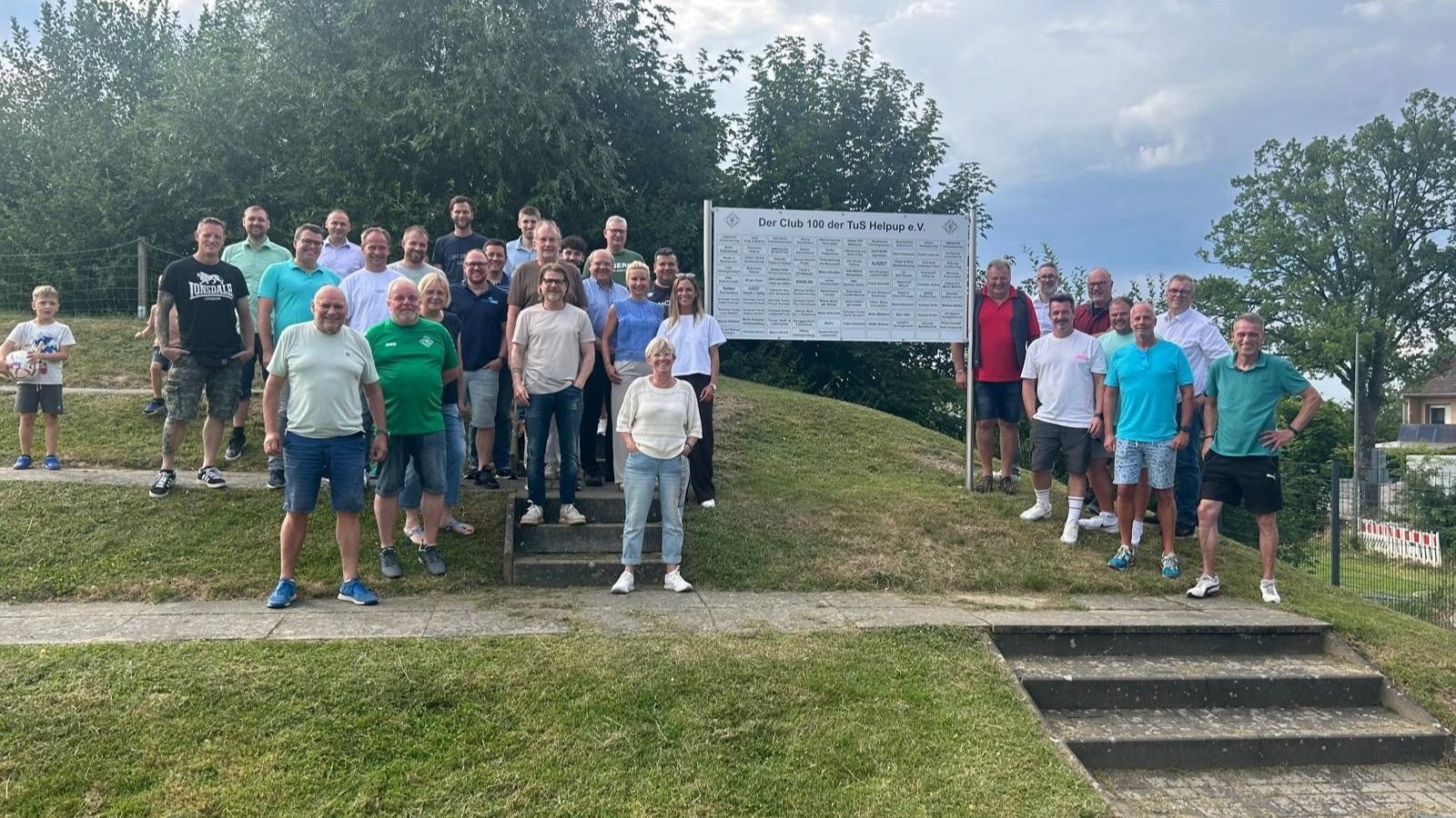 Eine große Gruppe von Männern und Frauen posiert mit einem Jungen und Fußball vor dem Schild "Club 100 der TuS Helpup