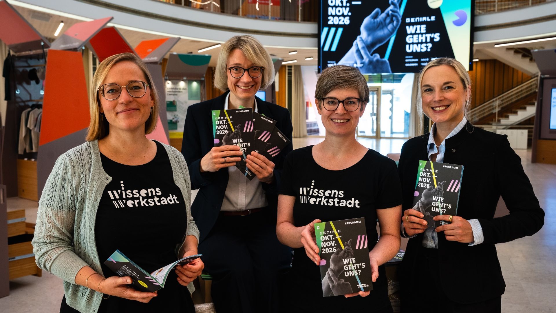 Vier lächelnde Frauen präsentieren in einem modernen Gebäude Programmhefte der GENIALE 2026 "Wie geht's uns?