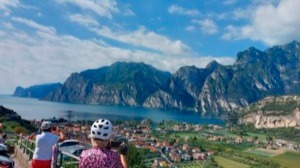 Zwei Personen mit weißen Helmen schauen auf einen großen See mit Bergen und einem Uferort unter blauem Himmel