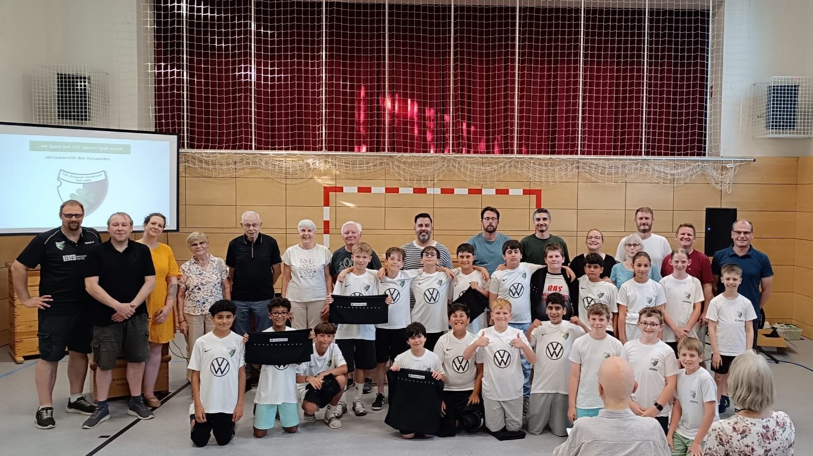 Viele Erwachsene und Kinder einer Sportmannschaft posieren in einer Sporthalle vor einer Projektion und einem