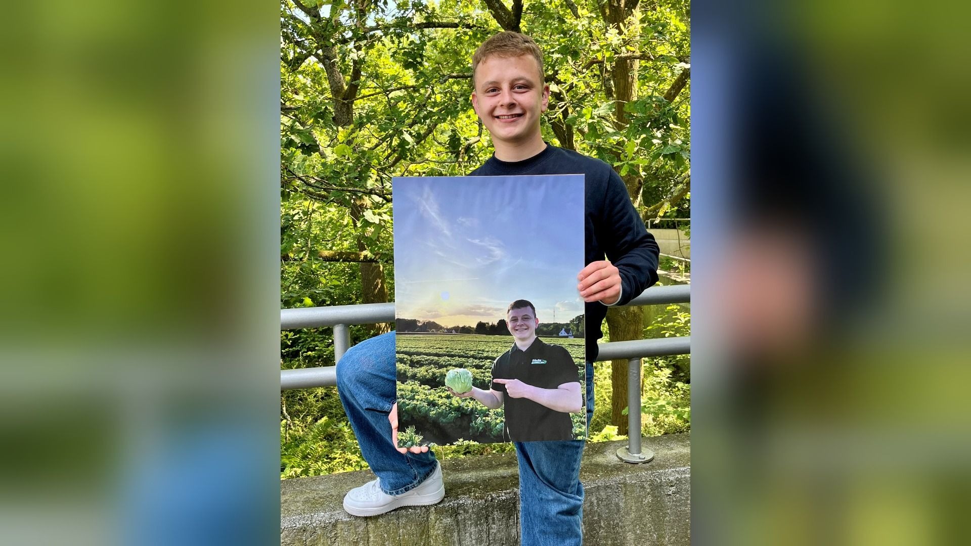 Junger Mann hält lächelnd ein großes Foto von sich mit einem Salatkopf im Feld bei Sonnenuntergang, vor grünem Laub