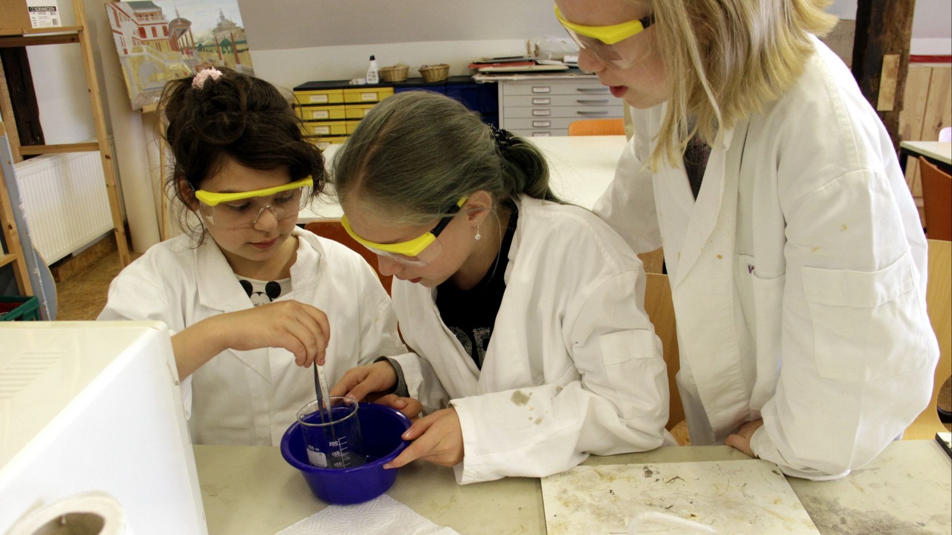 Drei Kinder in Laborkitteln und Schutzbrillen rühren mit einer Zange in einem Becherglas in einer blauen Schüssel
