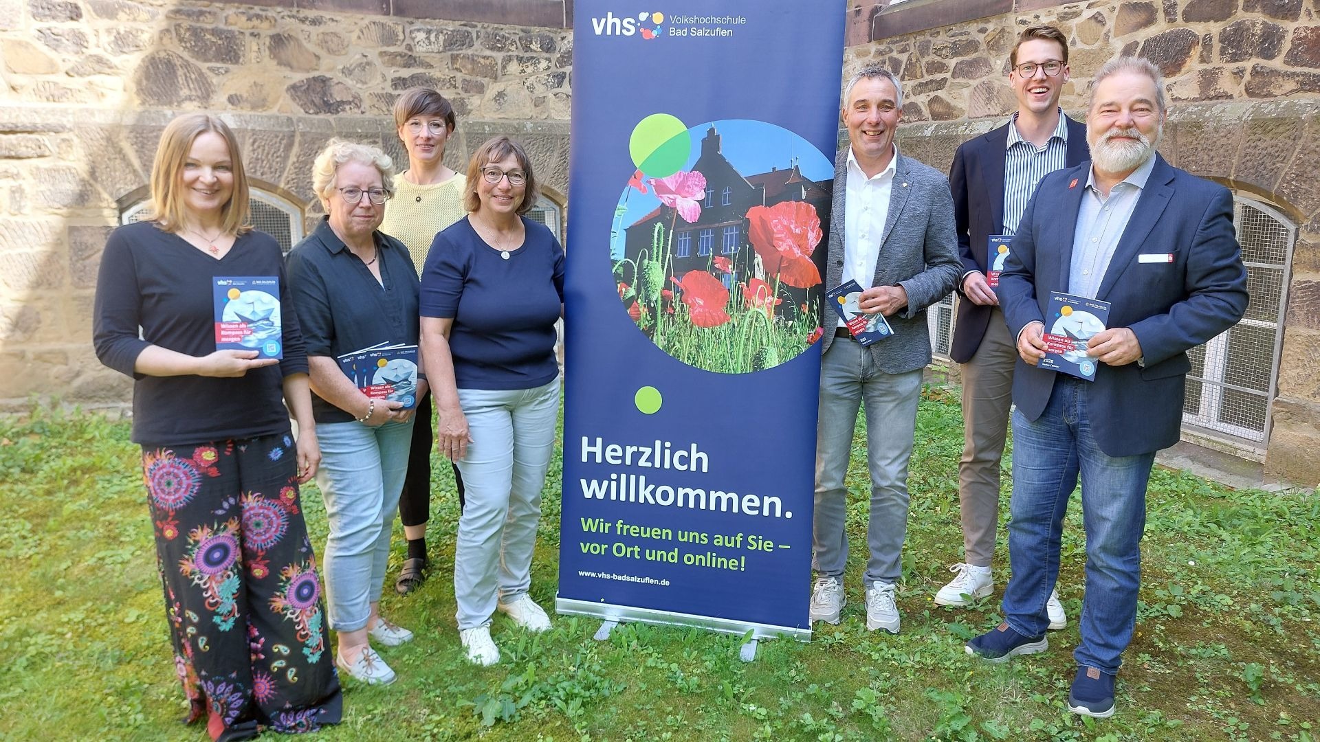 Sieben lächelnde Personen stehen vor einem vhs-Banner und einer Steinwand, halten Broschüren "Wissen als Kompass für