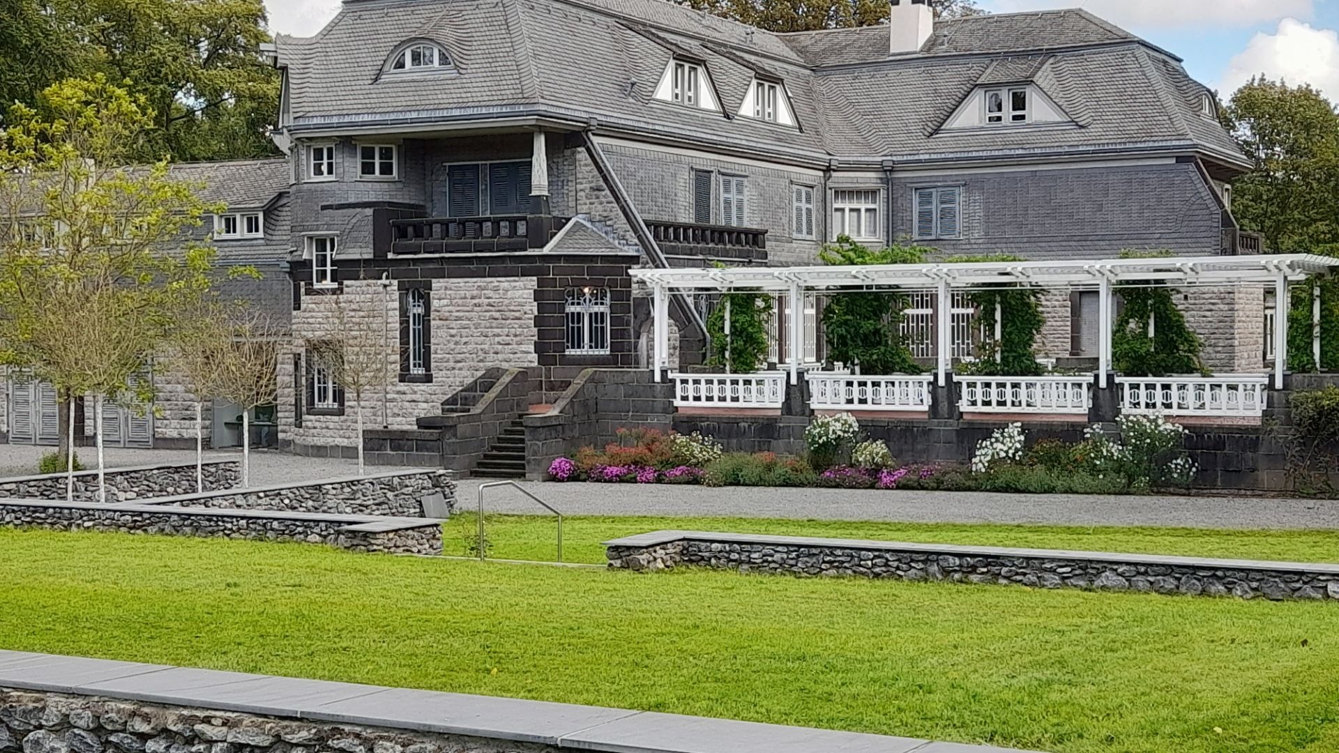 Graue Jugendstilvilla mit Schieferdach, weißer Pergola und Blumenbeeten in einem Garten