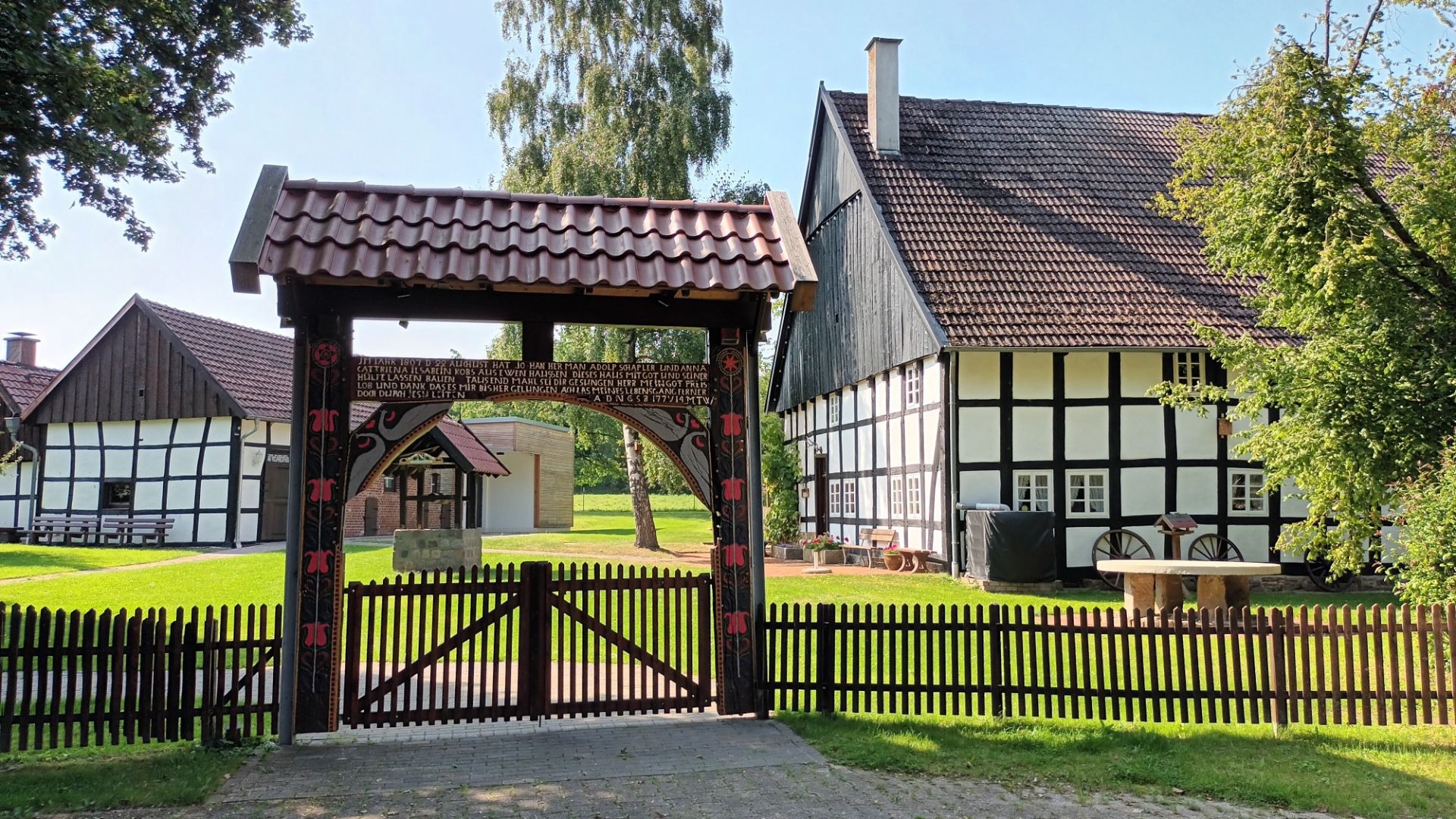Verzierter Holzbogen mit Inschrift als Eingang zu historischen Fachwerkhäusern mit gepflegtem Garten