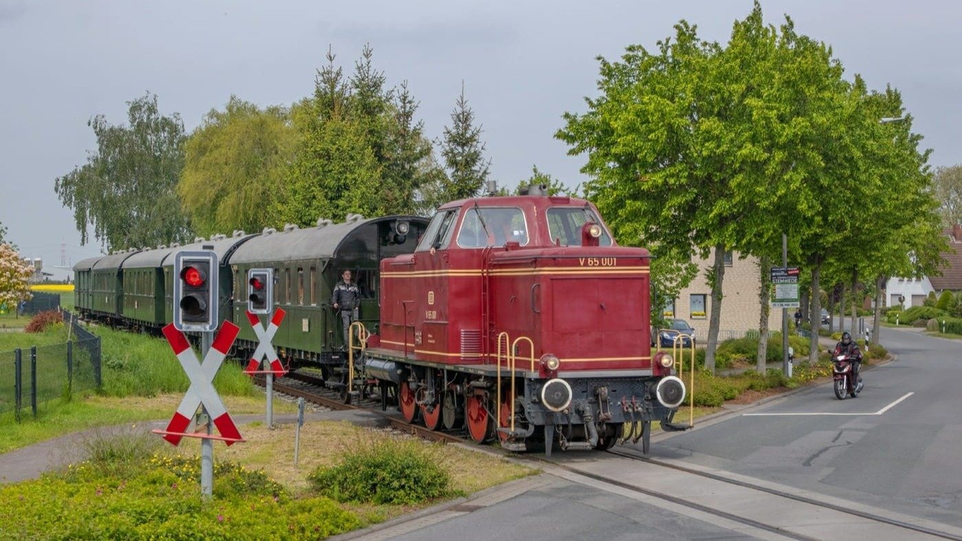 Historische rote Diesellok mit grünen Waggons an Bahnübergang mit roten Signalleuchten und einem Motorradfahrer auf der