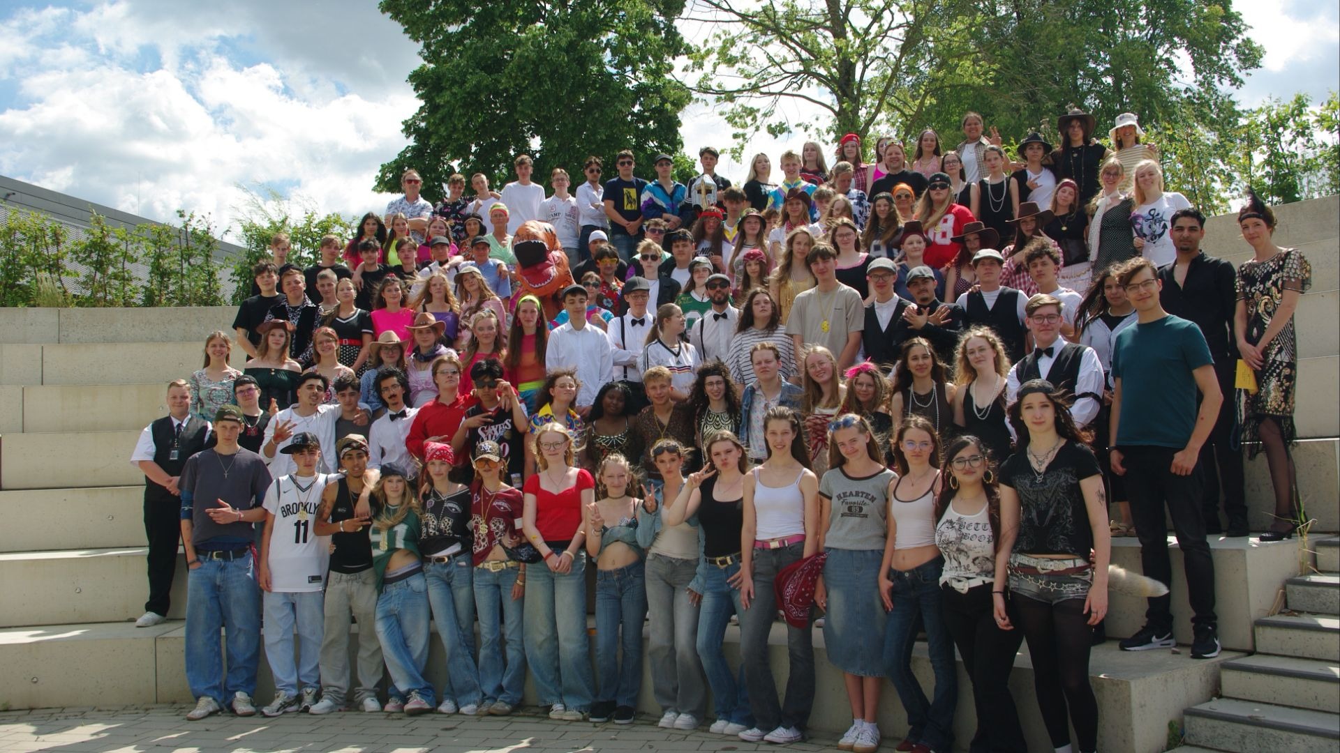 Eine große Gruppe Schüler in bunten Kostümen posiert auf Außentreppen vor Bäumen und blauem Himmel