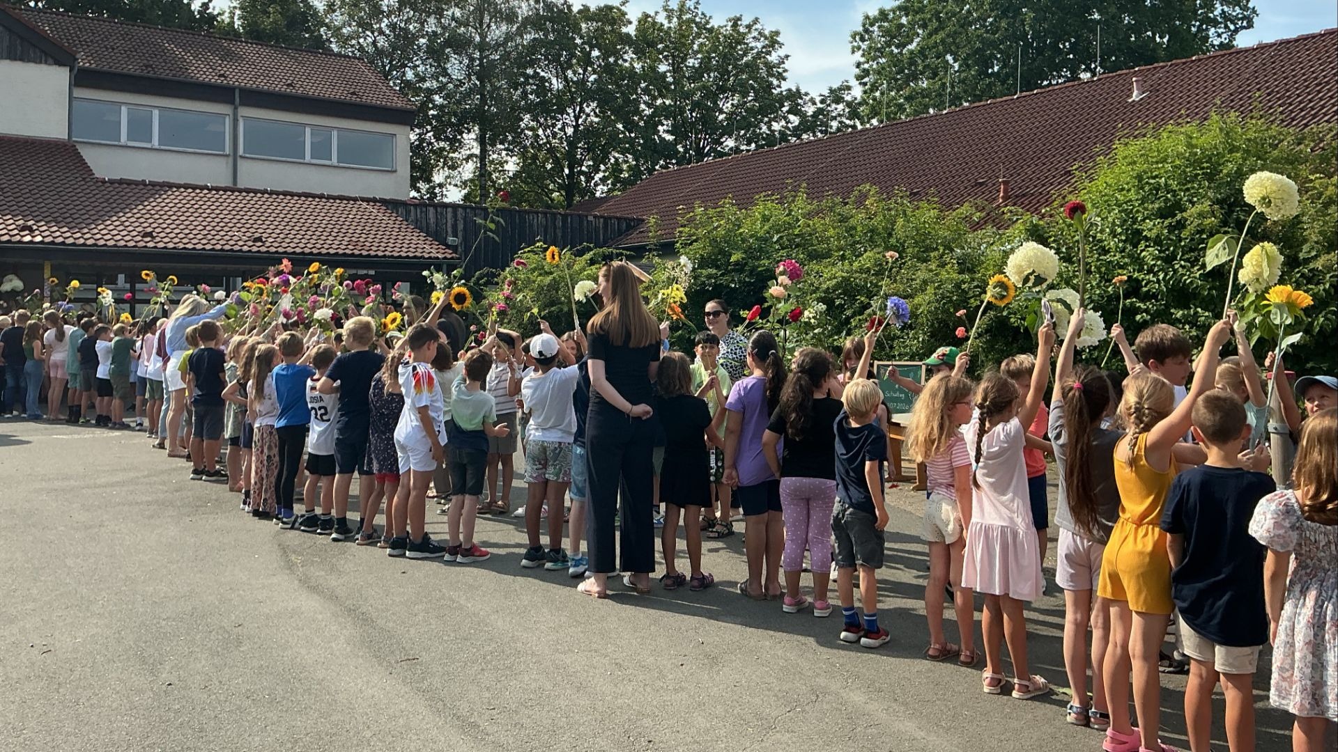 Kinder und Erwachsene stehen in einem langen Blumen-Spalier auf dem Schulhof vor einem Schulgebäude