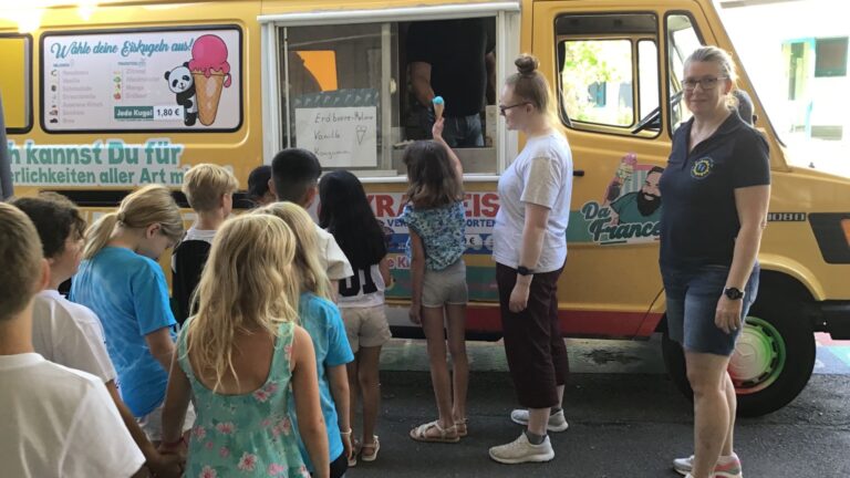 Personen, darunter viele Kinder, stehen vor einem gelben Eiswagen, ein Kind erhält eine Eistüte