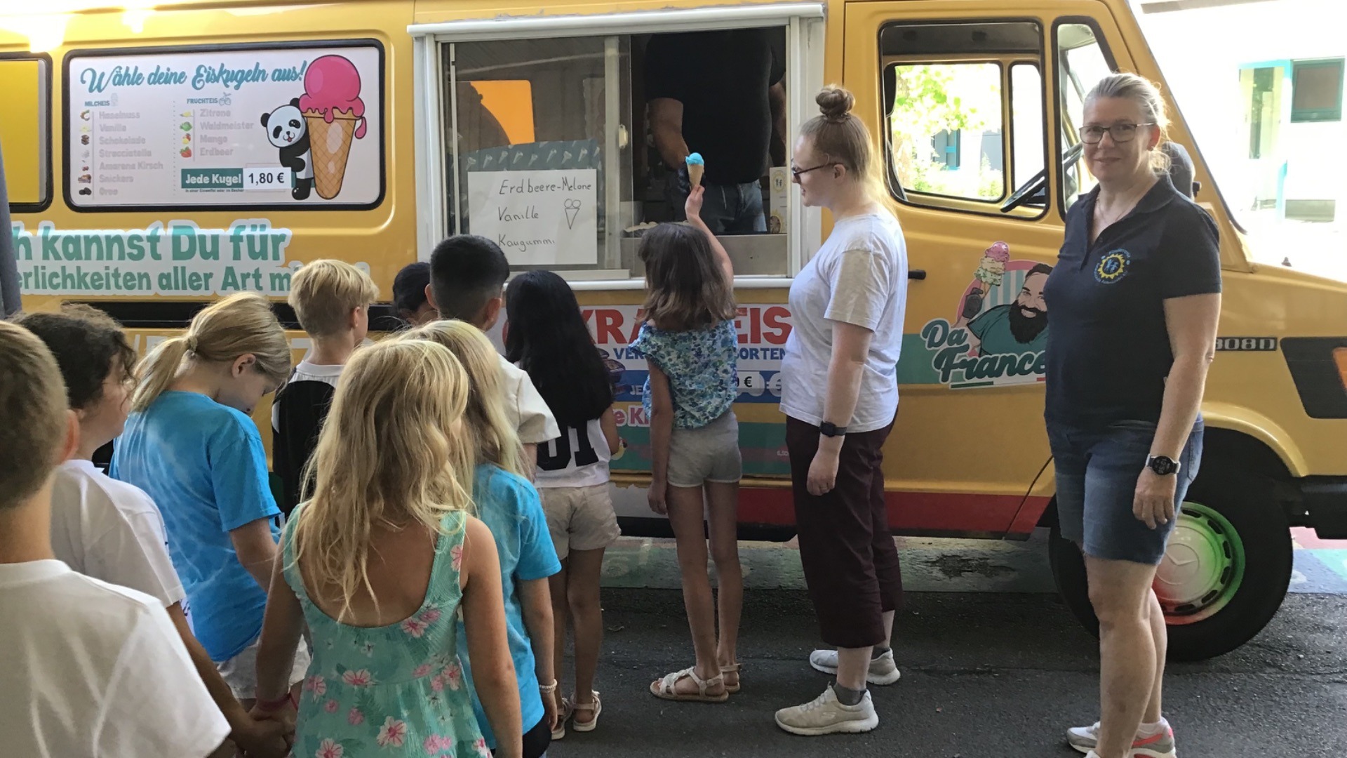 Personen, darunter viele Kinder, stehen vor einem gelben Eiswagen, ein Kind erhält eine Eistüte