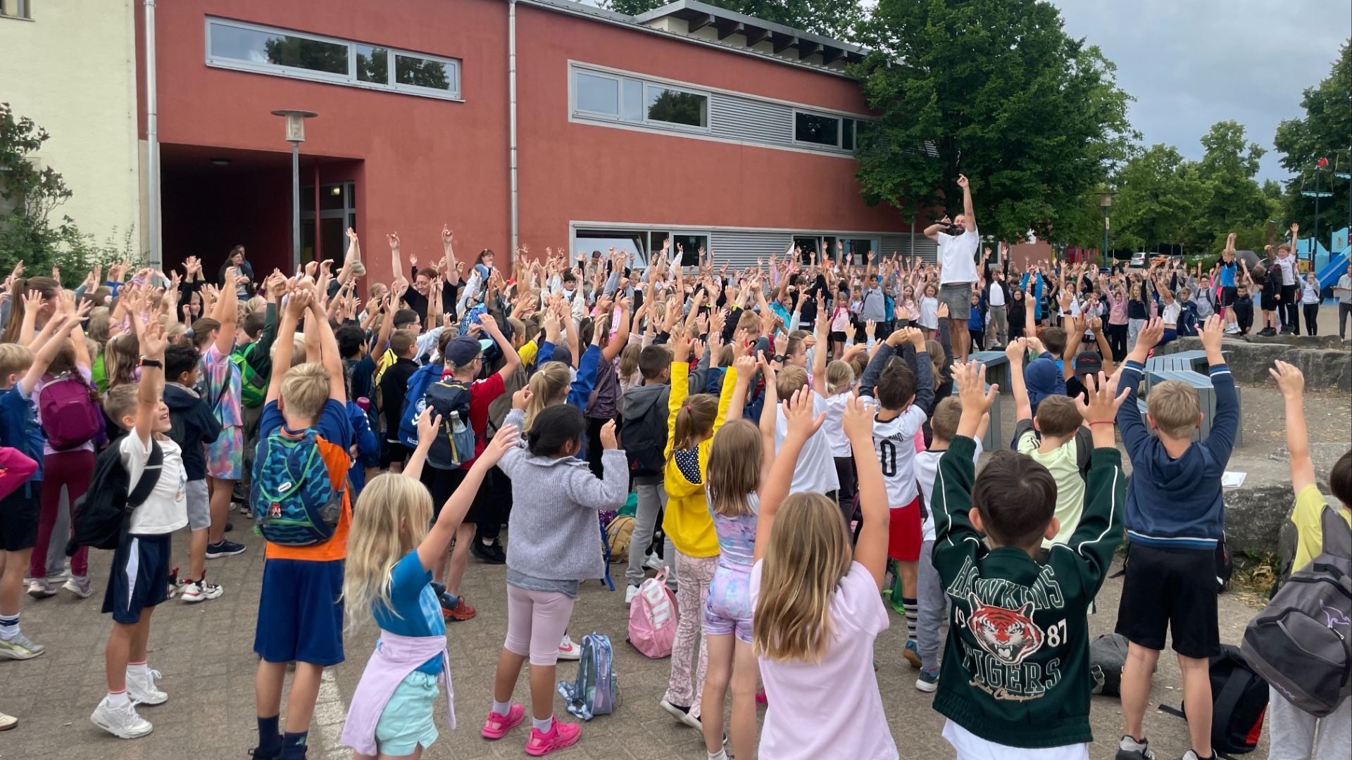 Viele Schulkinder strecken auf dem Schulhof ihre Arme in die Luft, angeleitet von einem Erwachsenen vor einem Gebäude