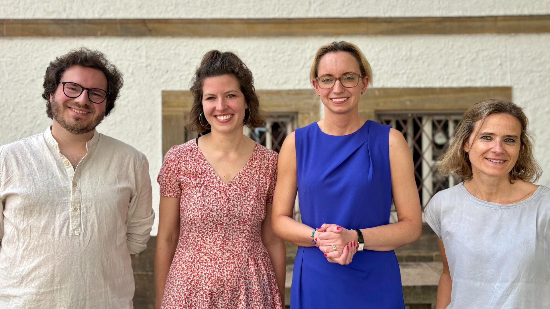 Vier lächelnde Personen, drei Frauen und ein Mann, stehen nebeneinander vor einem hellen Gebäude