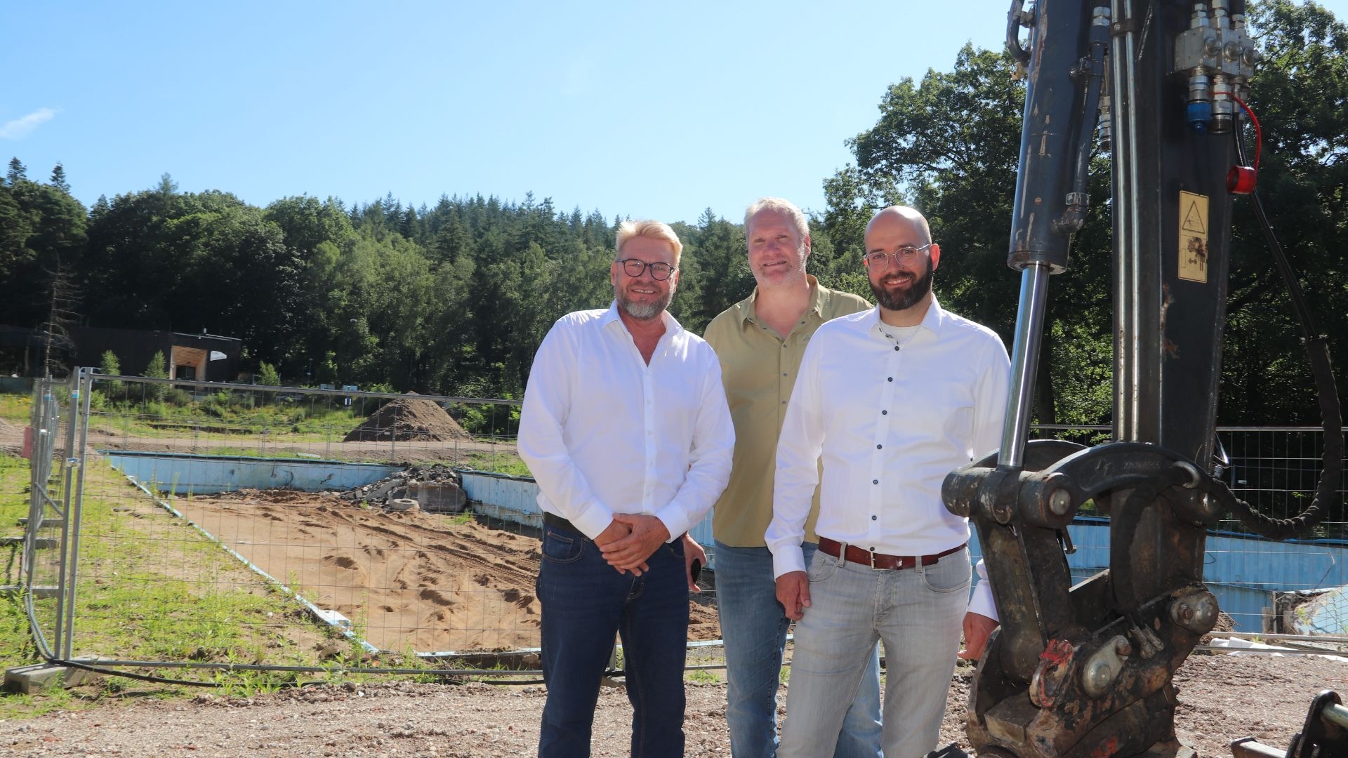 Drei Männer lächeln auf der Baustelle des Oerlinghauser Freibads mit abgerissenem Becken und Wald