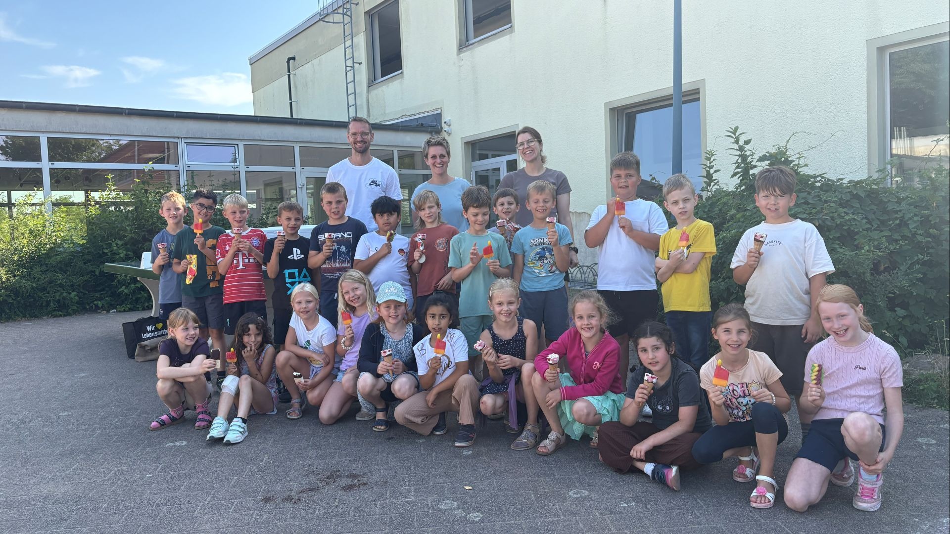 Rund dreißig lächelnde Schulkinder und drei Erwachsene stehen oder hocken mit Eis vor einem Schulgebäude