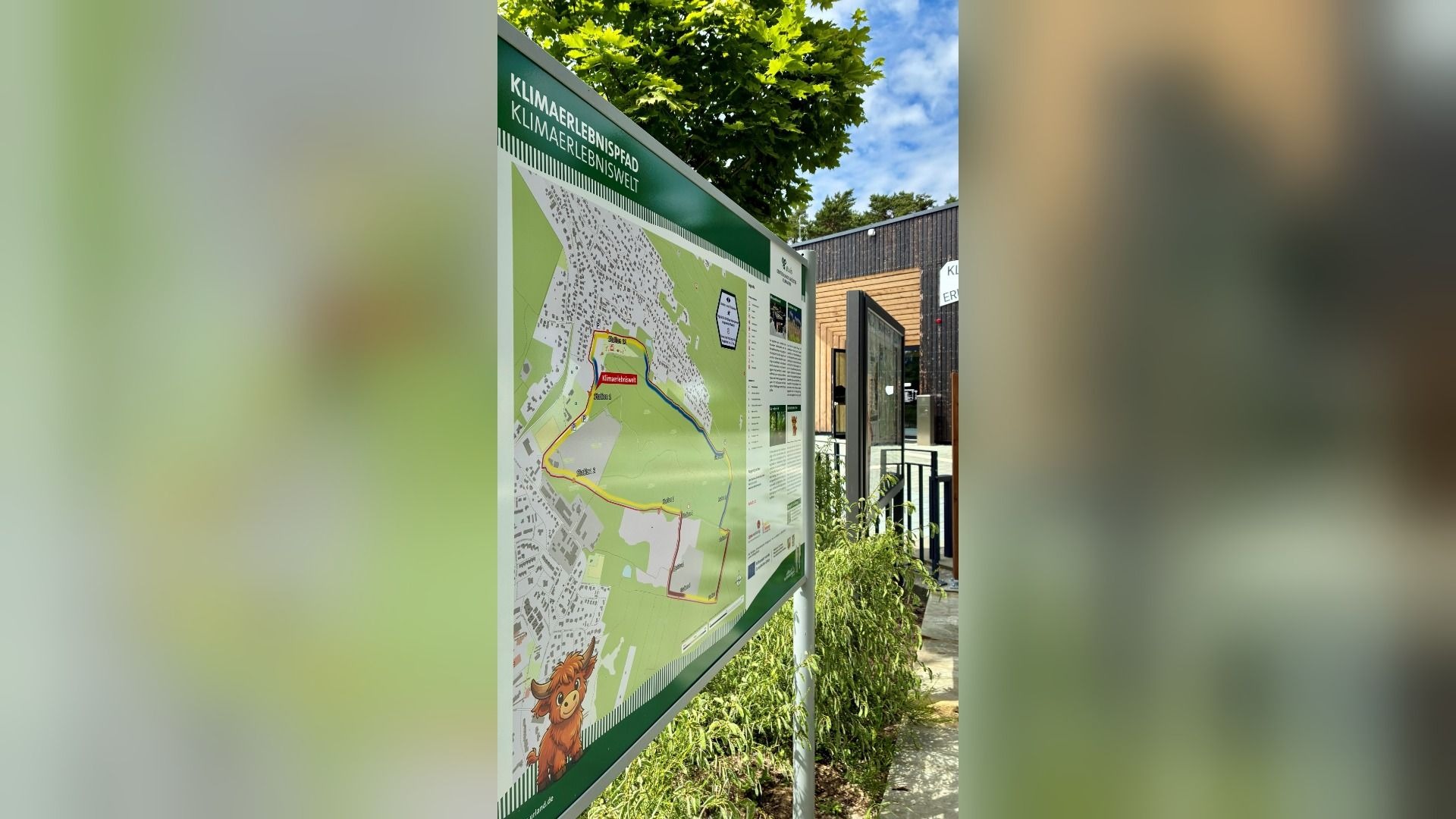 Grüne Informationstafel des Klimaerlebnispfades mit Karte des Weges und elf Stationen vor einem Holzgebäude