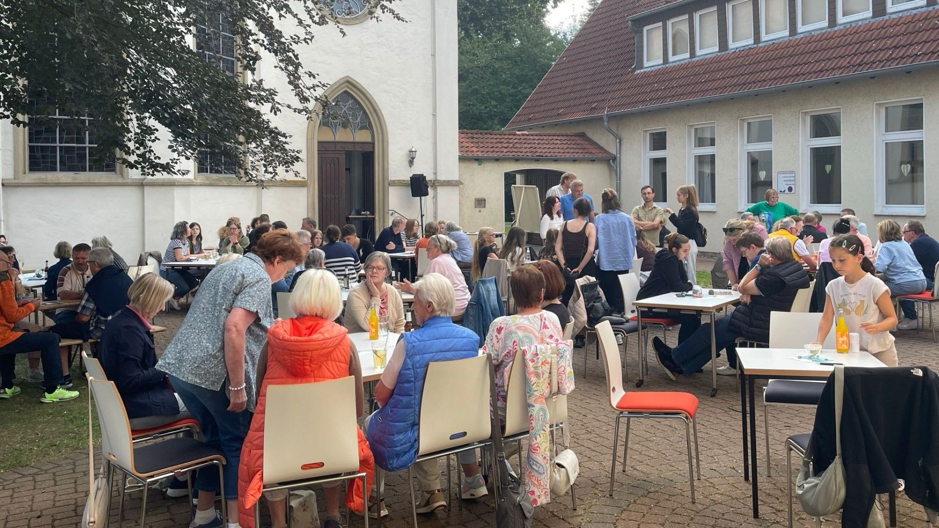 Eine große Gruppe Menschen sitzt und steht im Freien an Tischen zwischen einem weißen Kirchengebäude und einem Haus