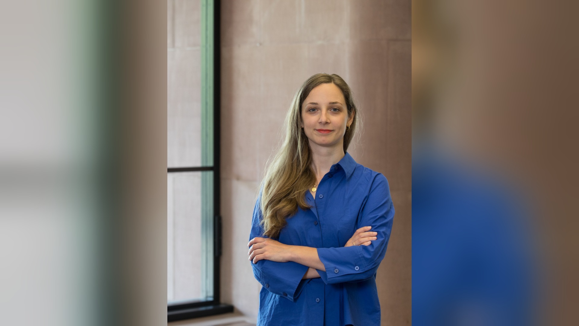 Kuratorin Laura Rehme steht mit verschränkten Armen und einem Lächeln vor einer hellbraunen Wand
