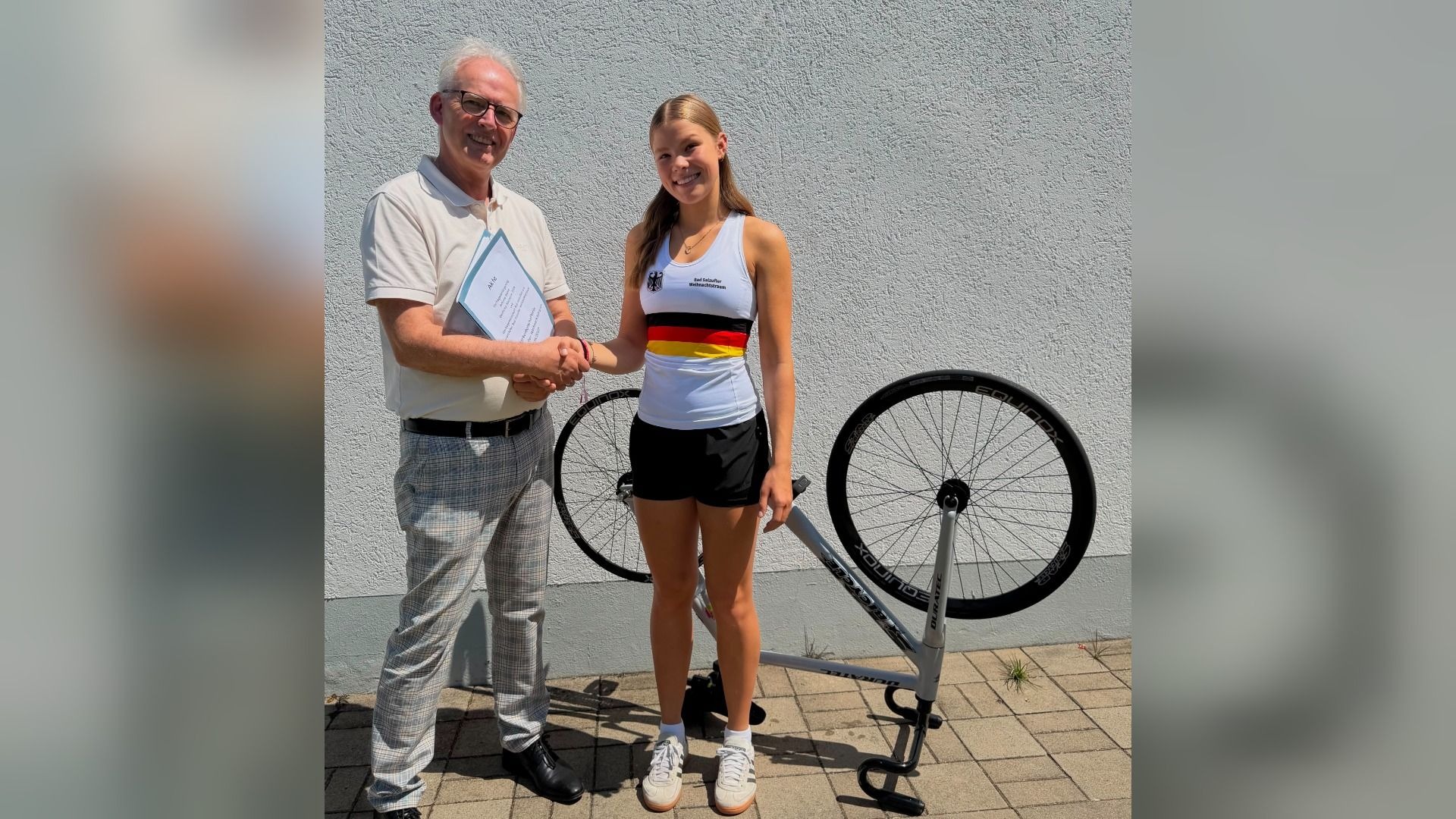 Antonia Blome im Deutschlandtrikot schüttelt Hände mit einem Mann vor einer weißen Wand und einem Rennrad auf Ständer