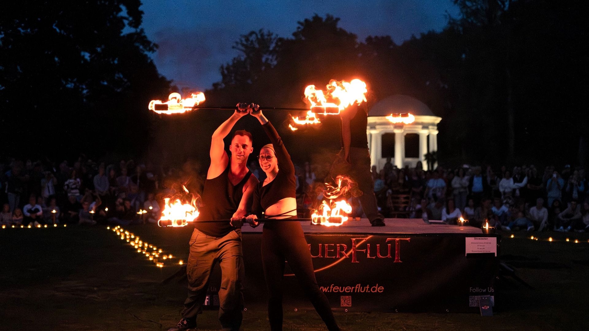 Zwei Personen mit brennenden Stäben und Kerzenlinien vor Zuschauern und einem Pavillon bei Nacht im Park