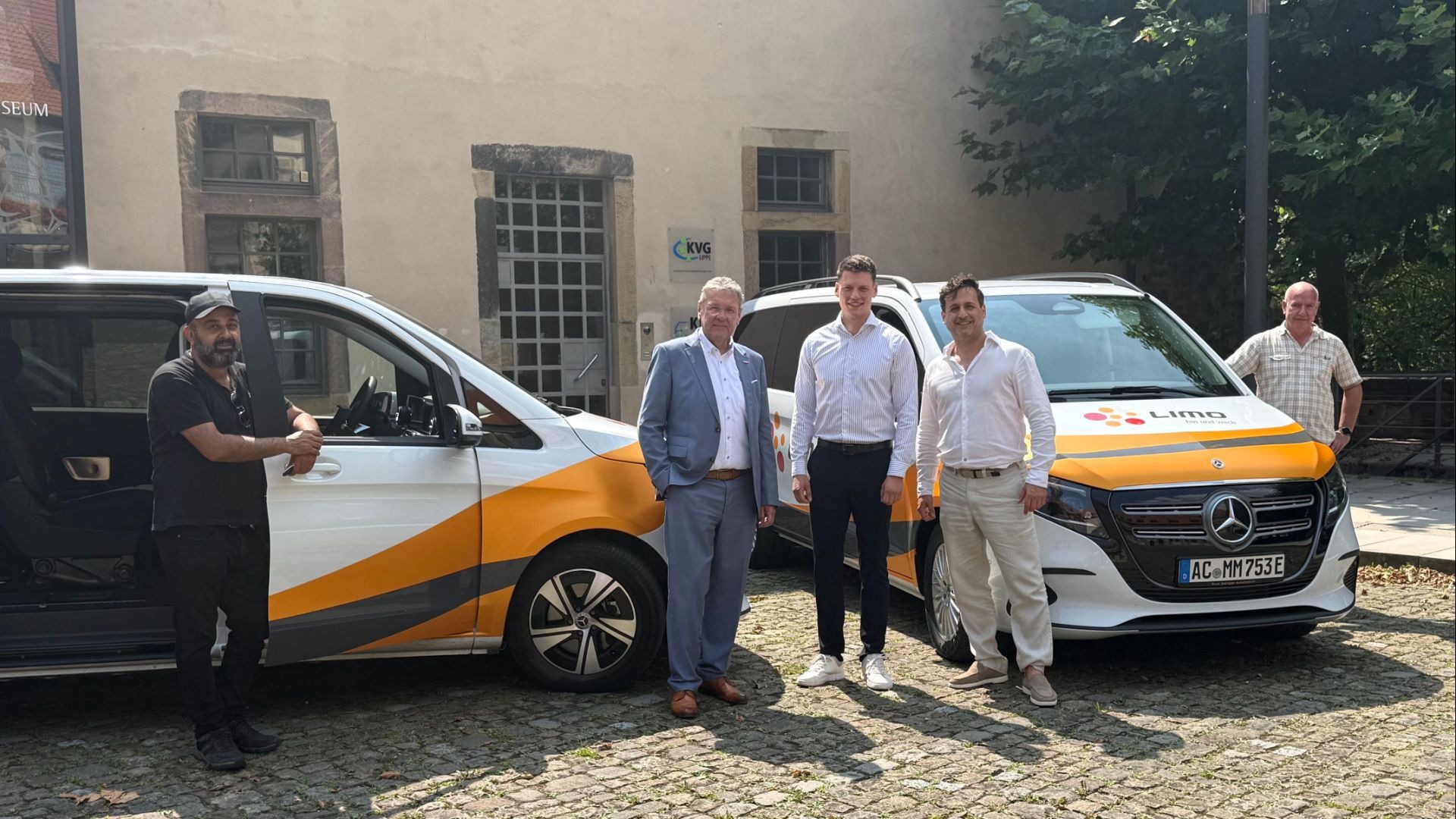 Fünf Männer posieren auf Kopfsteinpflaster vor zwei weißen LIMO-Vans mit orangefarbenen Streifen und dem Gebäude der