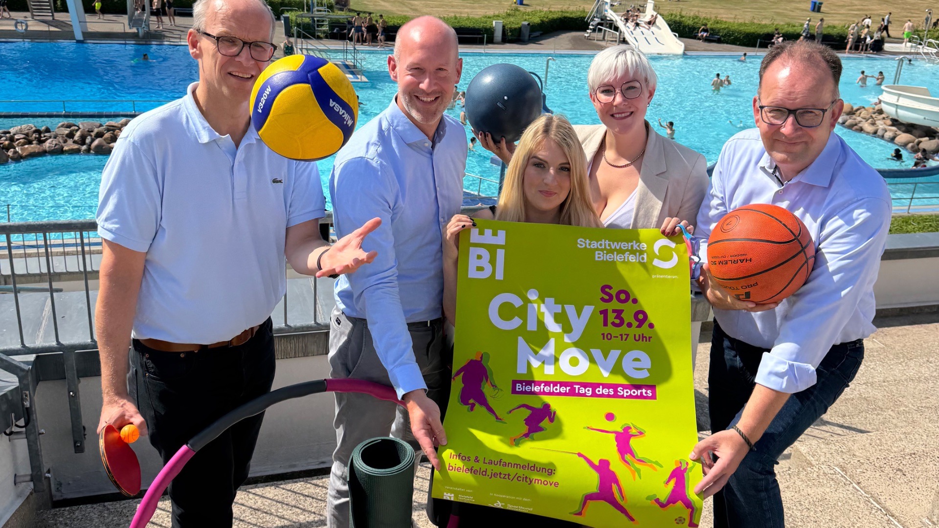 Fünf Personen halten ein grünes Plakat für den "City Move Bielefeld" und Sportgeräte an einem Schwimmbad