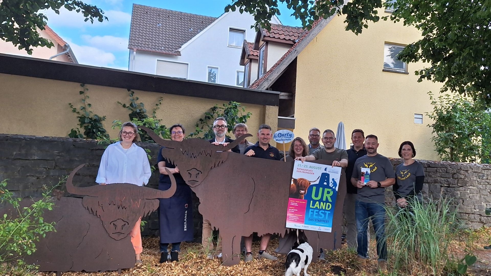 Eine Gruppe Personen, zwei große Metall-Kuhfiguren, ein Plakat für das UrLand-Fest und ein kleiner Hund stehen unter