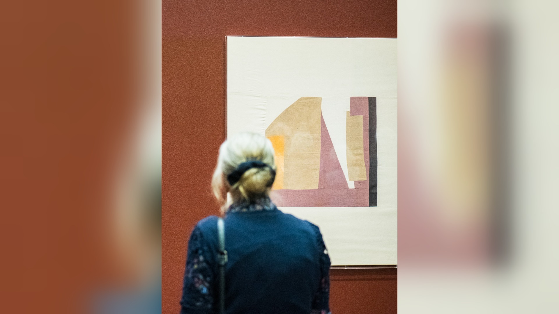 Eine Frau von hinten betrachtet ein abstraktes Kunstwerk mit geometrischen Formen an einer rotbraunen Wand in einer