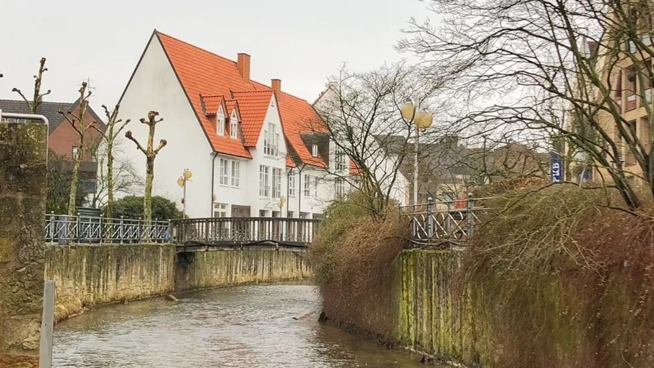 Fluss mit moosbewachsenen Ufern und Salze-Brücke, dahinter weiße Häuser mit roten Dächern in Bad Salzuflen