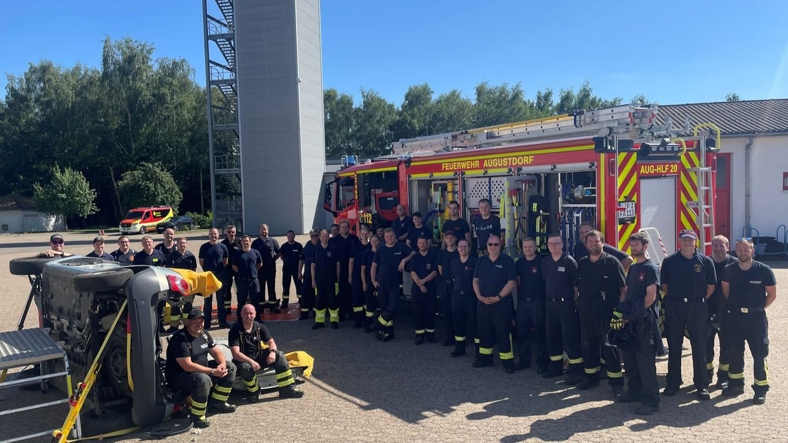 32 Feuerwehrleute aus Augustdorf posieren vor ihrem Löschfahrzeug und einem auf die Seite gelegten Auto