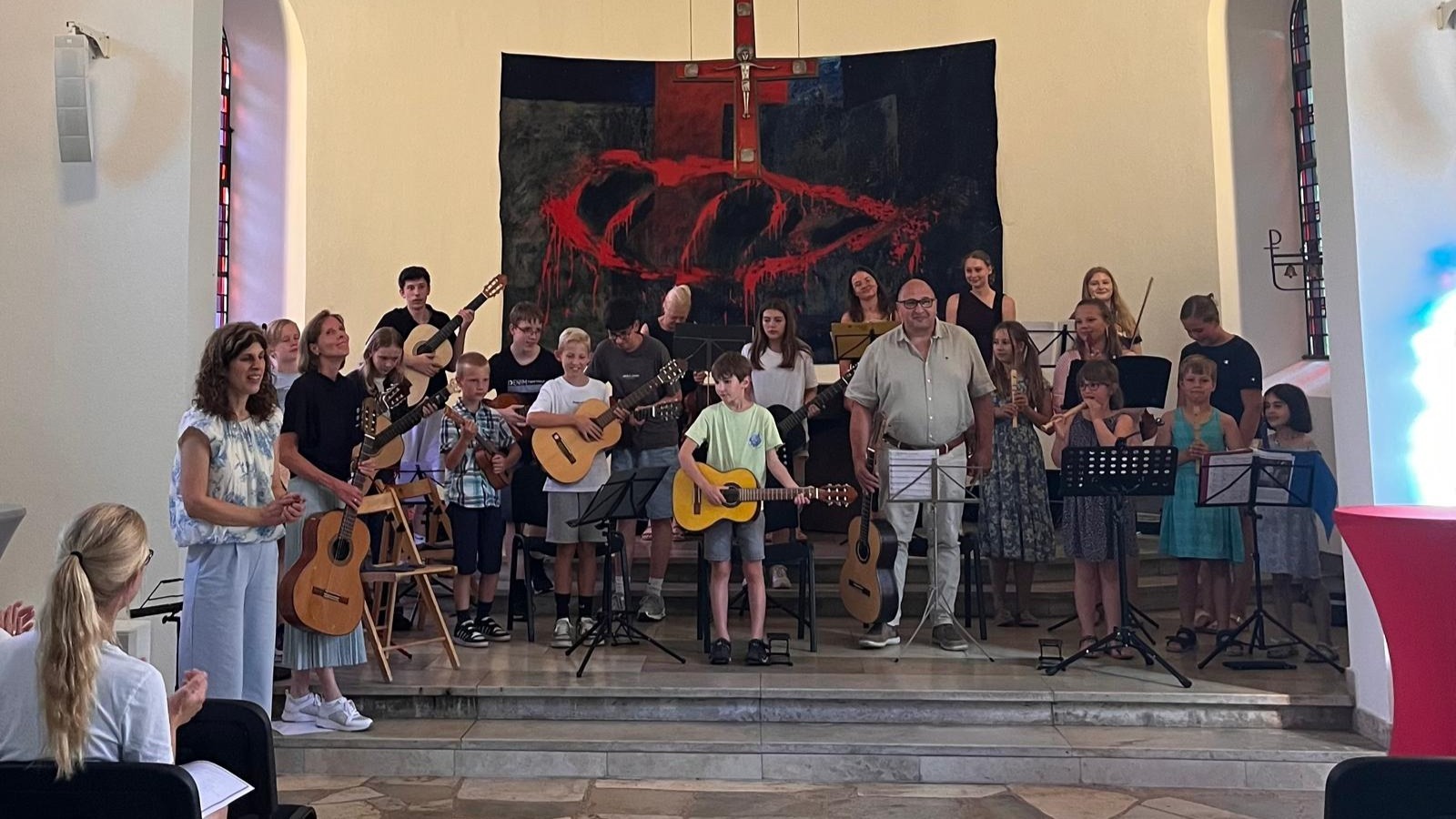Kinder und Erwachsene mit Gitarren, Flöten und Ukulele auf einer Bühne vor einem großen Kreuz in einer Kapelle