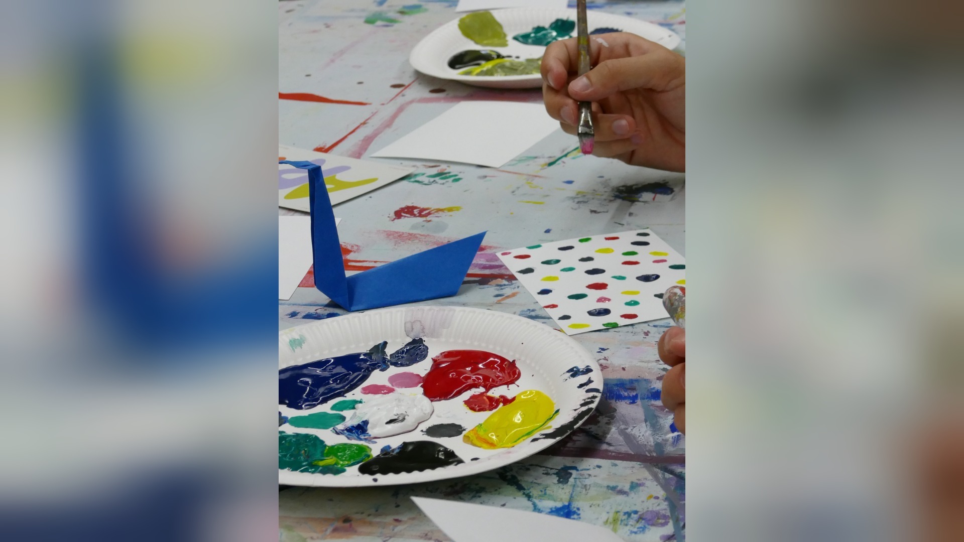 Hand hält Pinsel über Palette mit Farben, daneben ein blauer Origami-Vogel und bemalte Papiere auf Ateliertisch