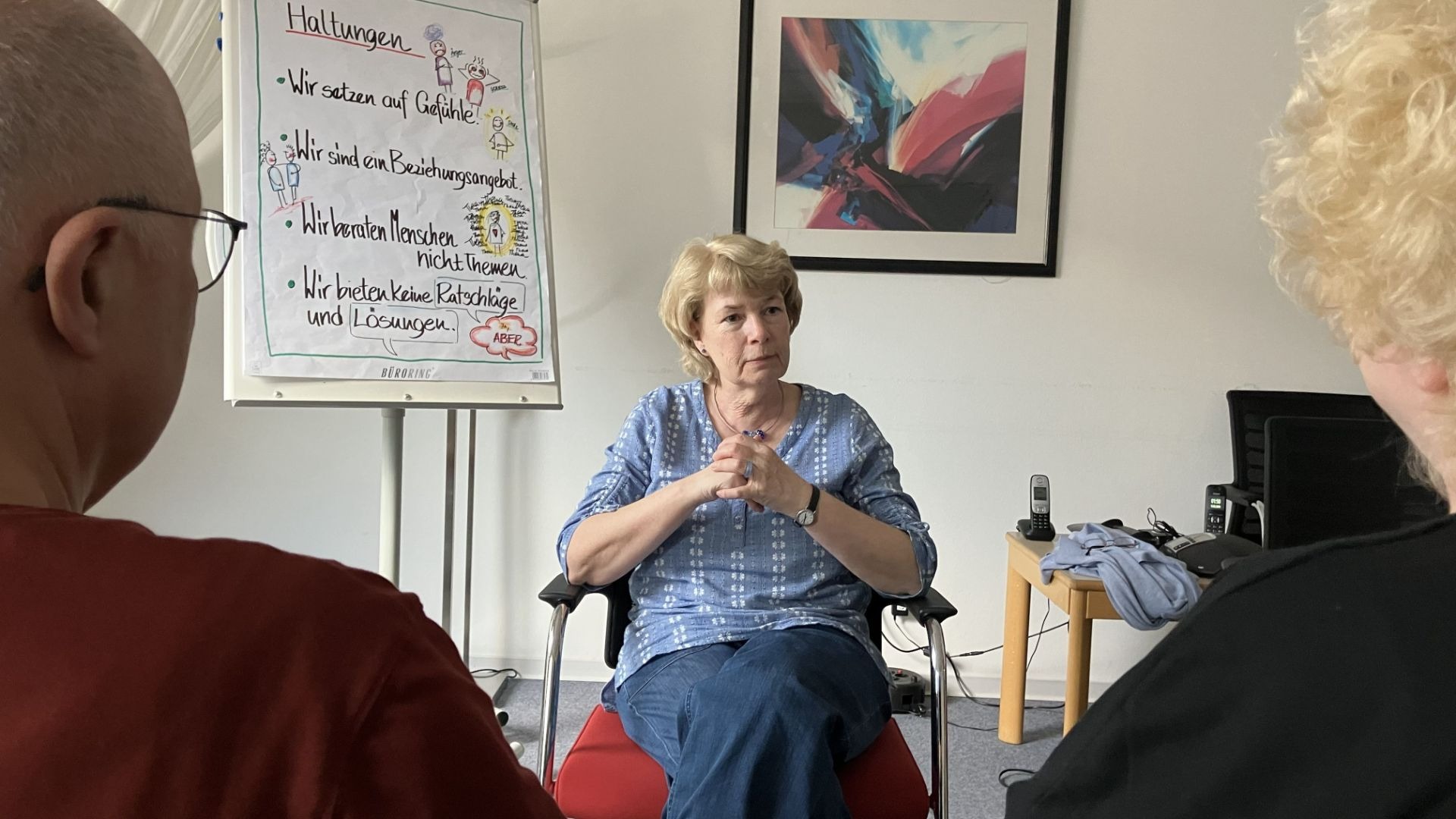 Drei Personen in einem Raum, eine Frau spricht, während zwei andere zuhören, hinter ihr ein Flipchart mit Überschrift