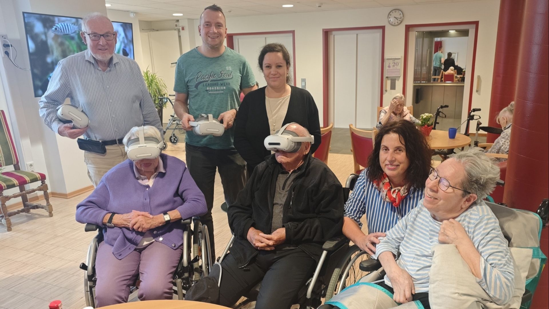 Zwei Senioren tragen VR-Brillen, umgeben von anderen Senioren und Betreuern in einem Aufenthaltsraum