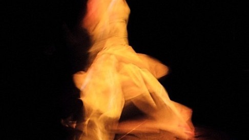 Eine in gelbem Licht dynamisch tanzende Person mit fließendem, hellem Kleid vor dunklem Hintergrund