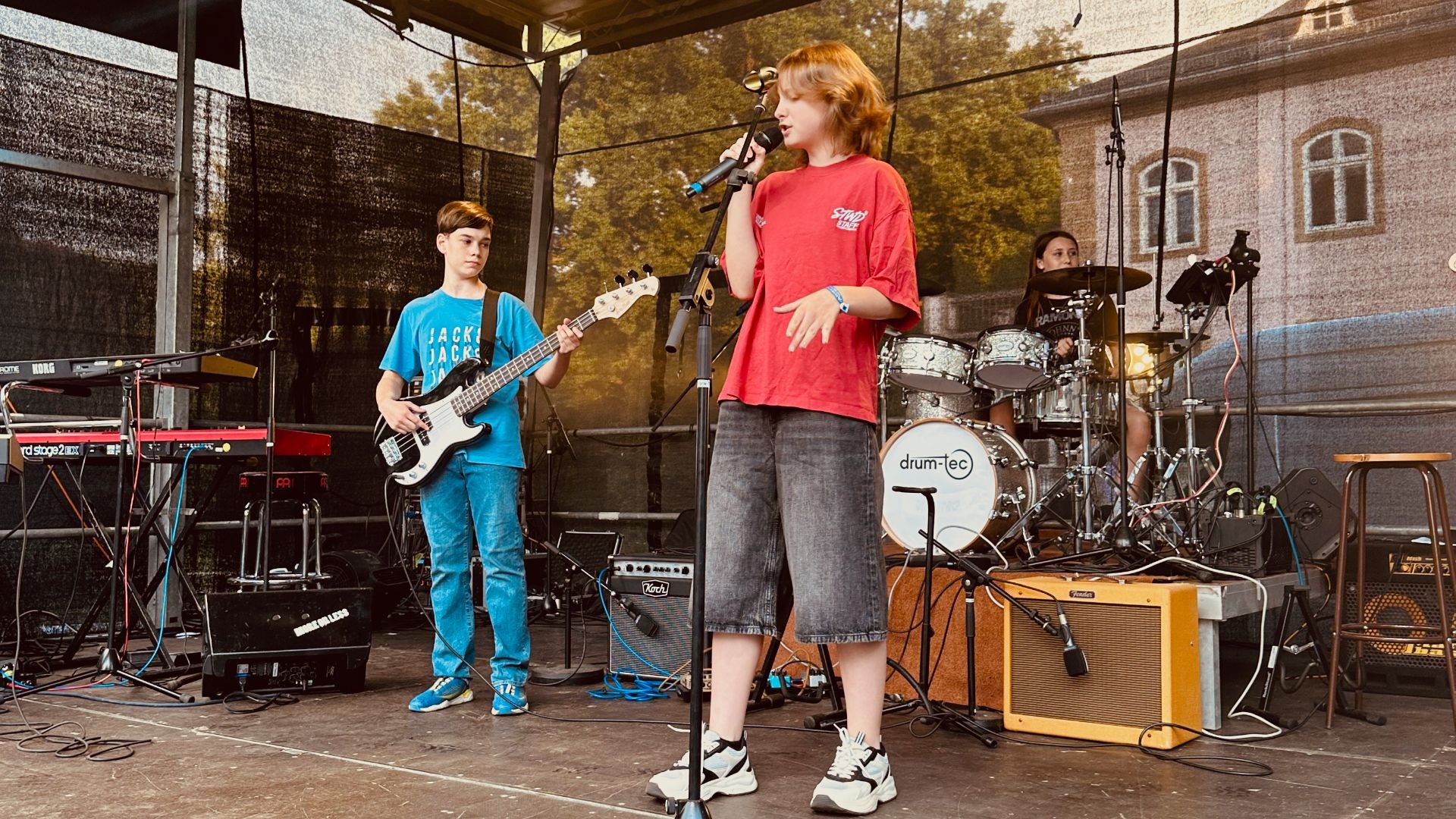 Eine junge Sängerin am Mikrofon, ein Bassist und eine Schlagzeugerin spielen auf einer Außenbühne