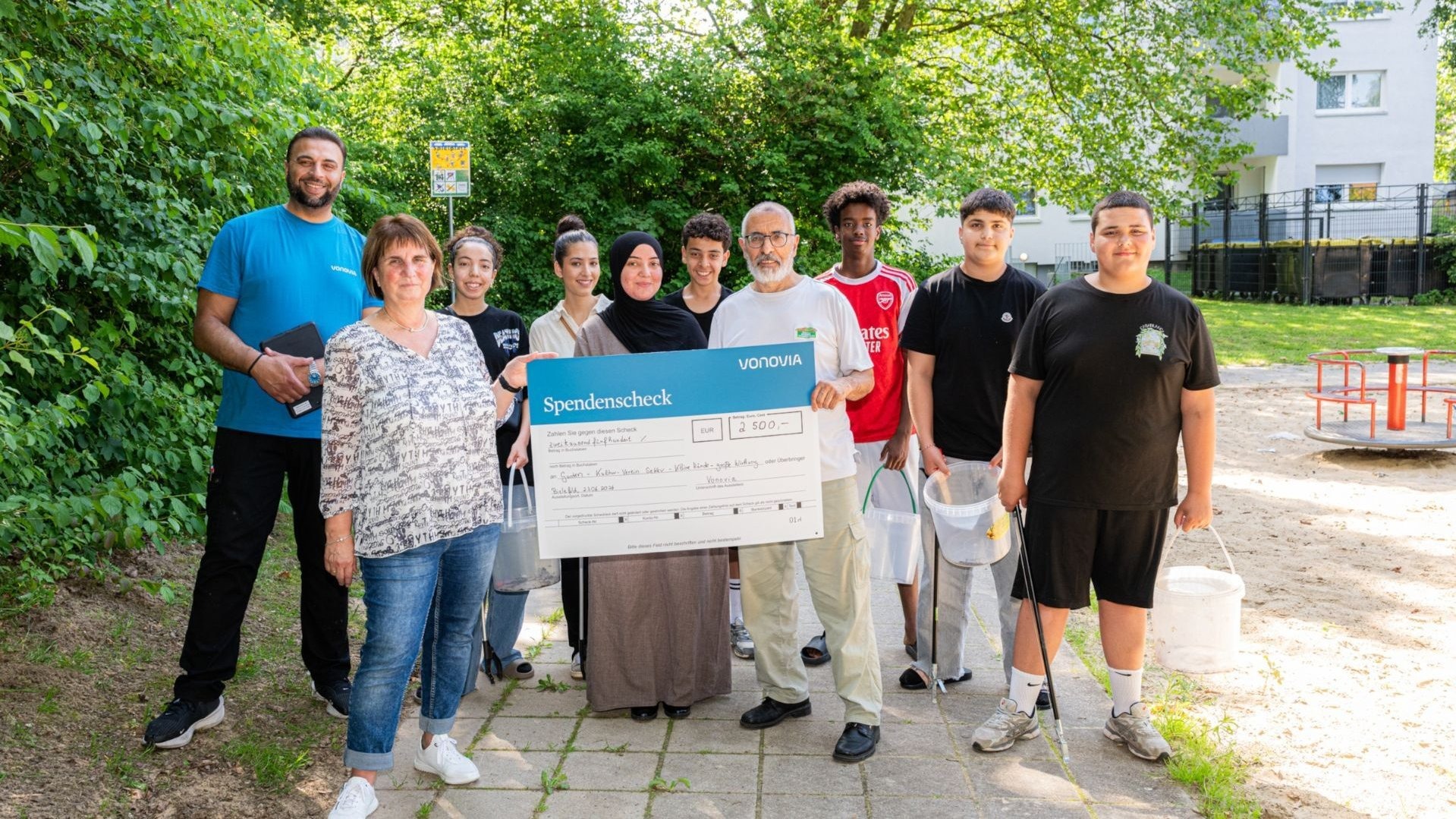 Eine Gruppe Erwachsener und Jugendlicher steht in Bielefeld im Park und präsentiert einen 2.500 Euro Spendenscheck von