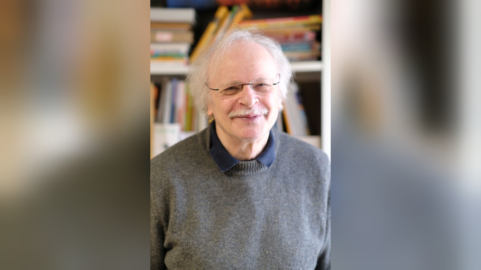 Ein lächelnder älterer Mann mit weißem Haar, Brille und Schnurrbart vor einem Bücherregal