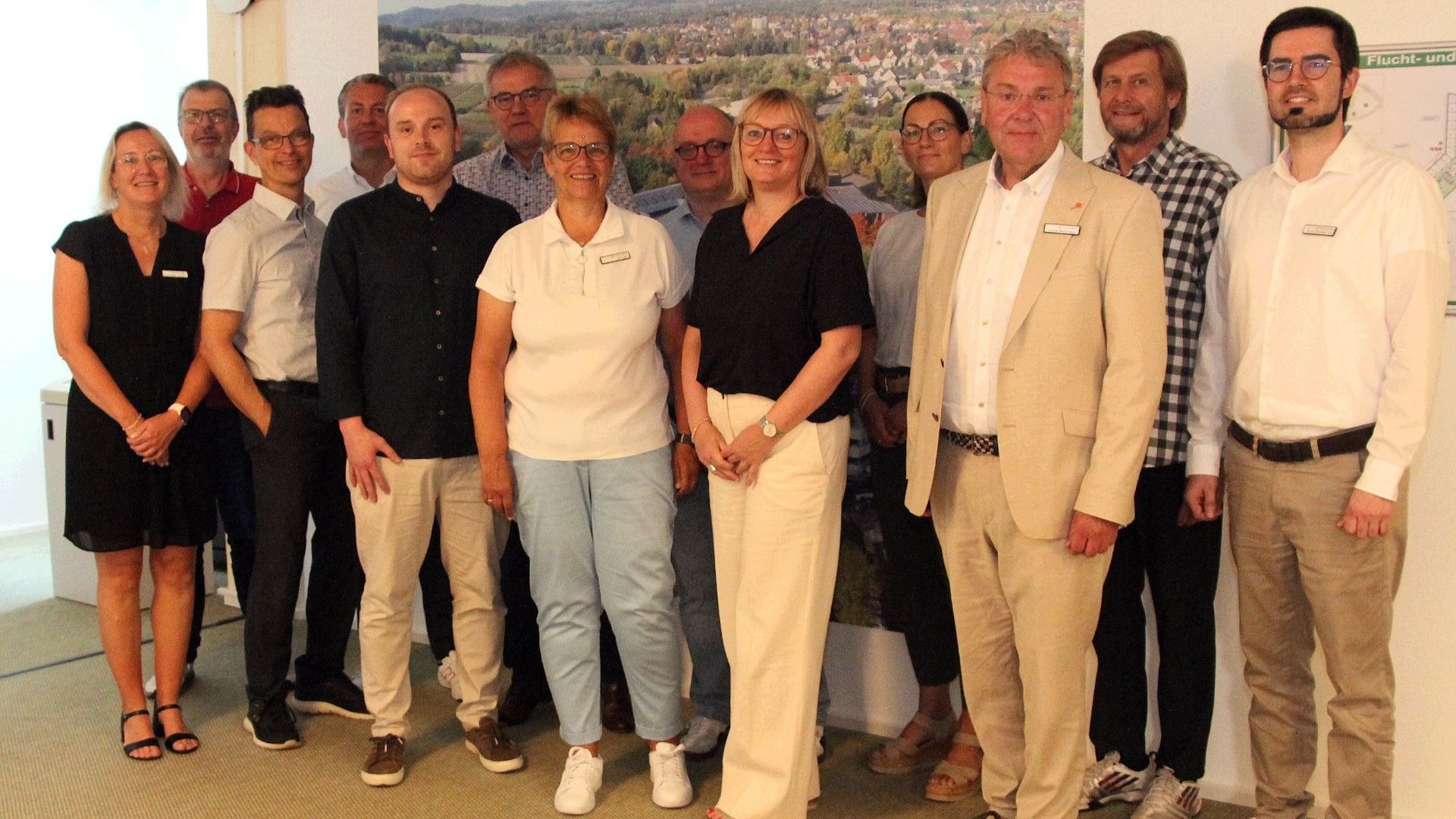 Elf lächelnde Männer und Frauen stehen in einem Raum vor einer großen Wandgrafik einer Siedlung