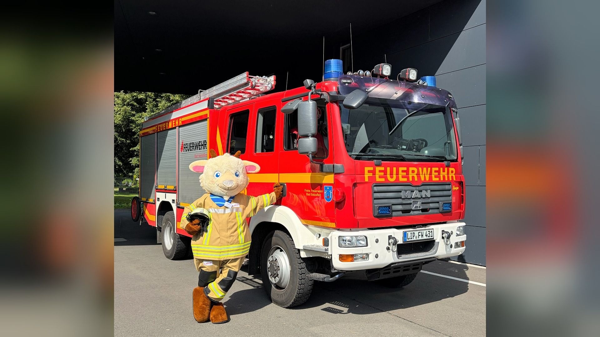 Stadtmaskottchen Ziege Line in Feuerwehruniform lehnt an einem roten Feuerwehrauto mit der Aufschrift FEUERWEHR