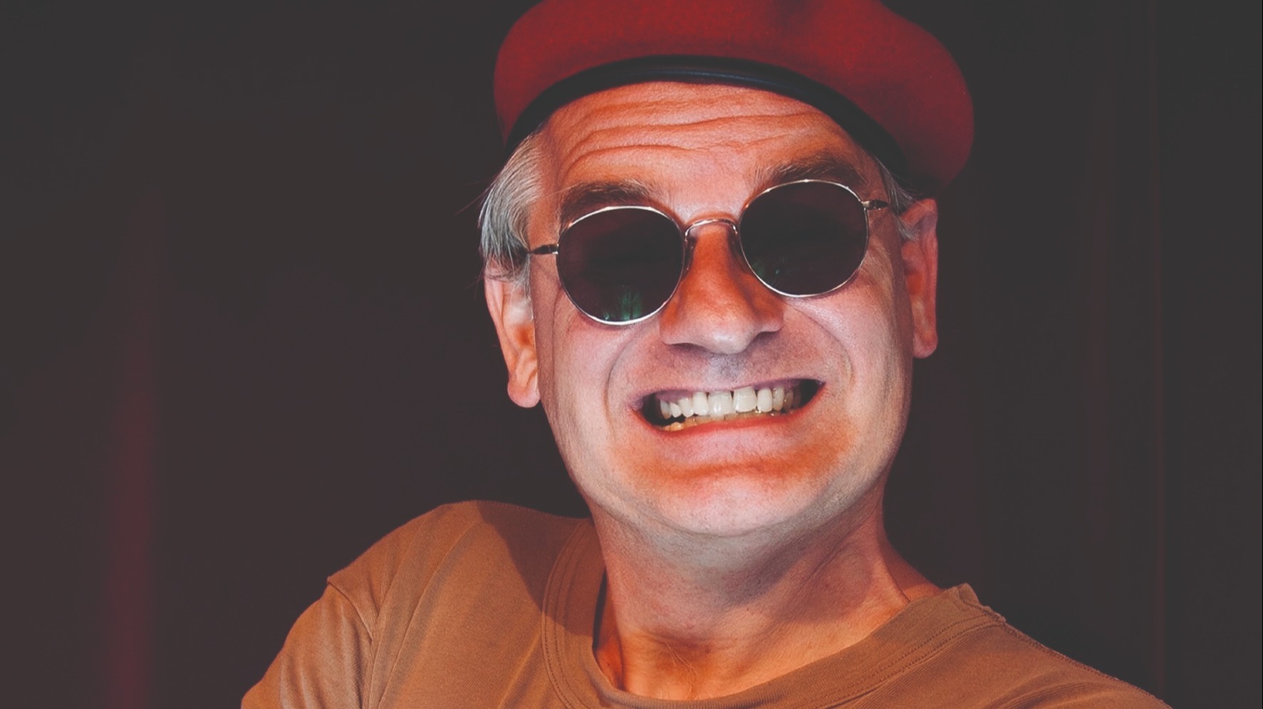 Mann mit rotem Barett und runder Sonnenbrille lächelt breit mit verschränkten Armen vor dunklem Hintergrund
