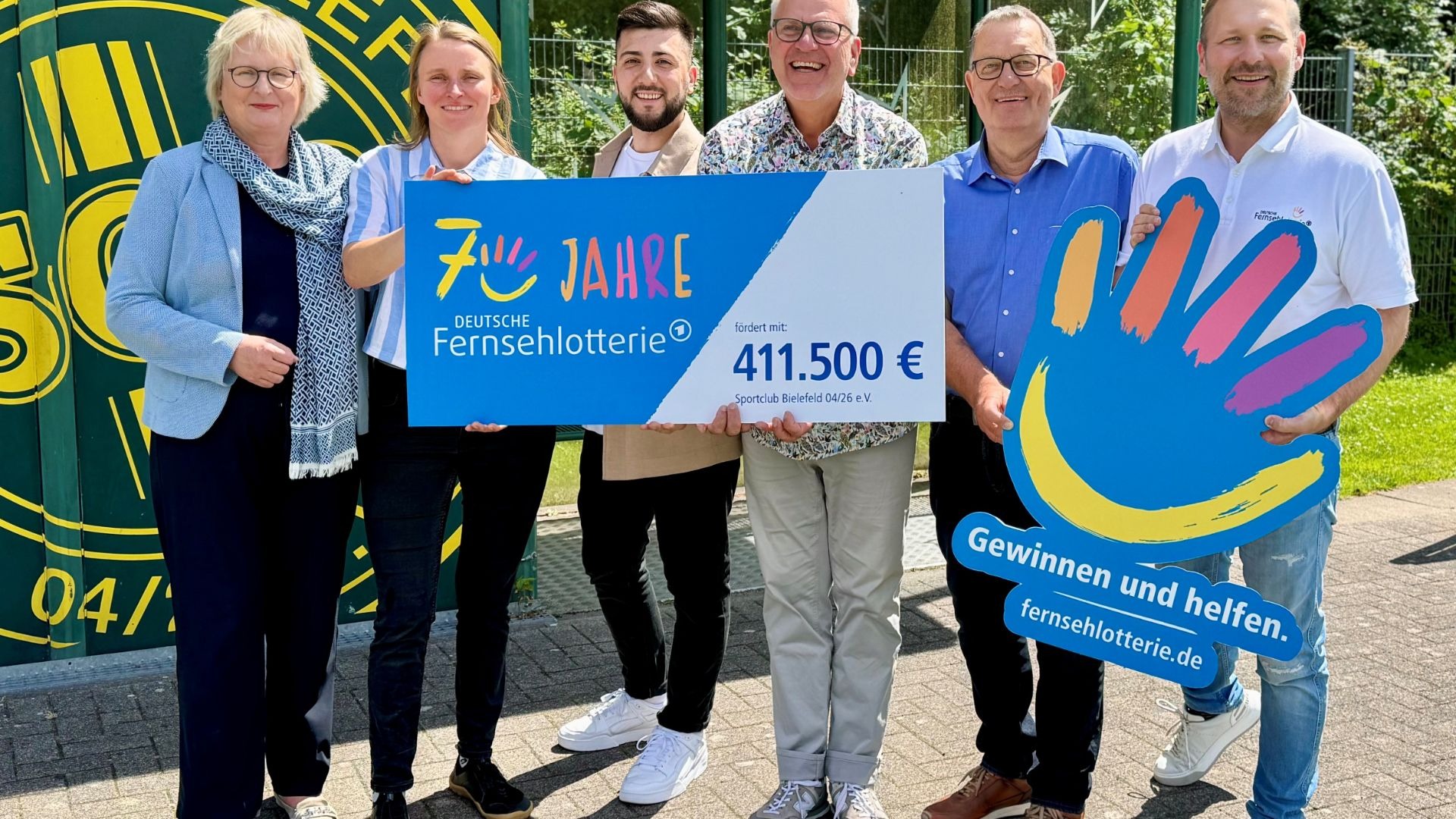 Sechs lachende Menschen halten einen großen blauen Scheck der Deutschen Fernsehlotterie über 411.500 Euro für Sportclub