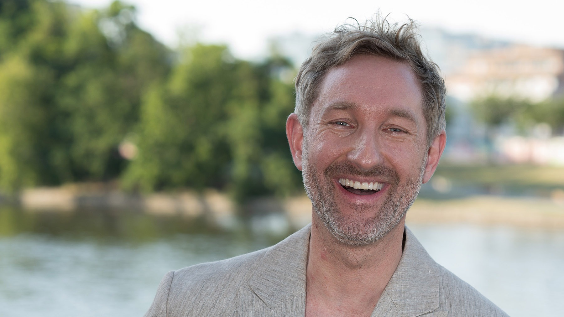Daniel Boschmann lacht breit, trägt ein helles Sakko, im Hintergrund Bäume und ein Fluss