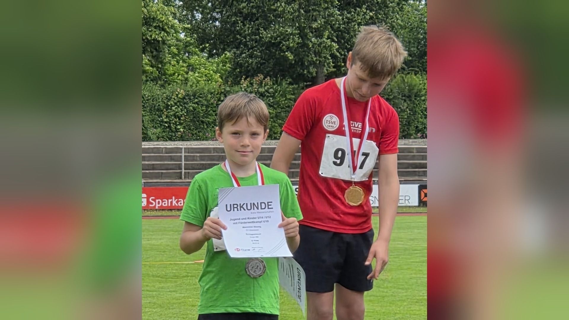 Zwei Jungen auf einem Podest, der linke mit grünem Shirt hält eine Urkunde und Silbermedaille, der rechte im roten
