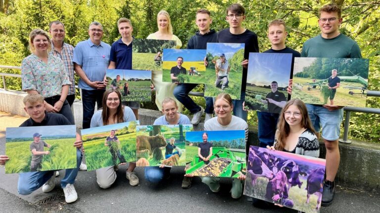 Fünfzehn Personen, viele junge Landwirte, halten draußen vor grünen Bäumen Plakate mit Landwirtschaftsmotiven