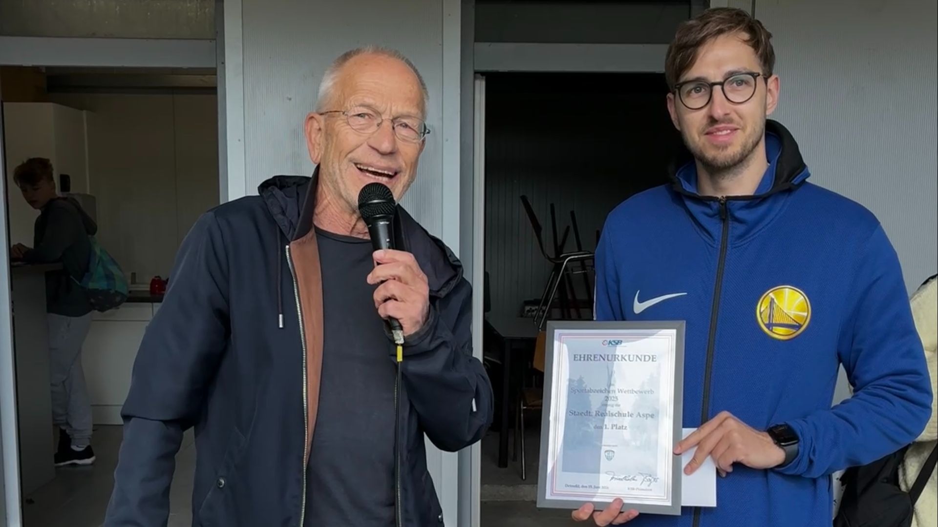 Ein älterer Mann mit Mikrofon und ein jüngerer Mann in blauer Jacke halten eine Ehrenurkunde für die Realschule Aspe