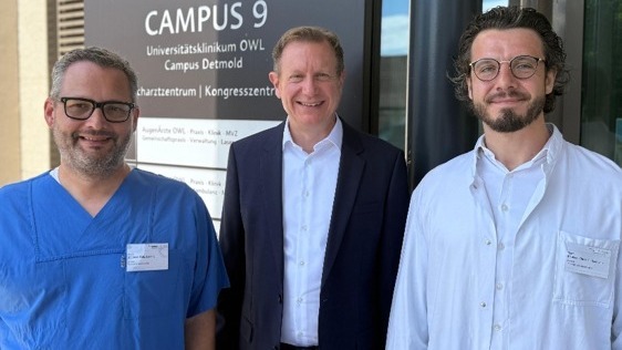 Drei lächelnde Männer vor dem Eingang des Universitätsklinikums Campus Detmold, zwei in medizinischer Kleidung, einer