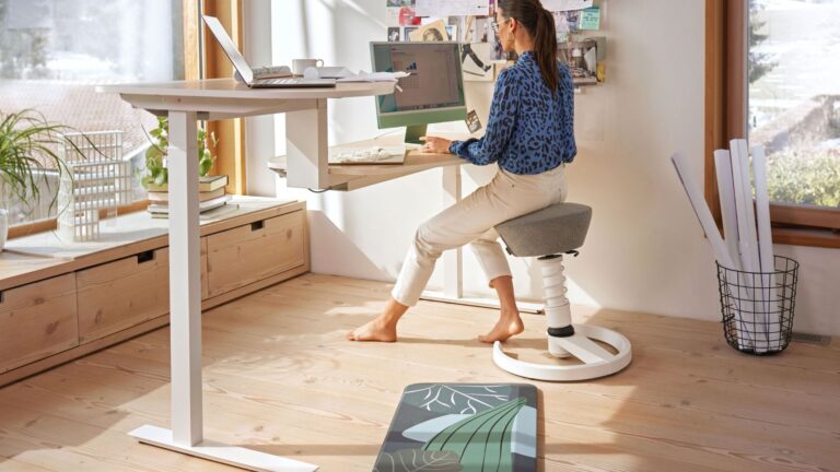 Frau arbeitet auf ergonomischem Hocker an einem Schreibtisch mit zwei Arbeitsplatten und Computern vor einem Fenster