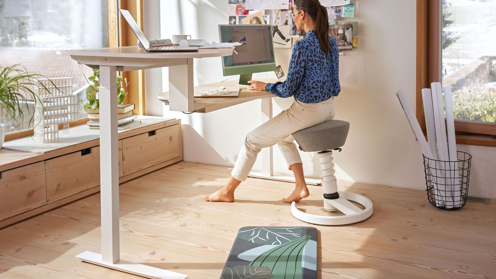 Frau arbeitet auf ergonomischem Hocker an einem Schreibtisch mit zwei Arbeitsplatten und Computern vor einem Fenster