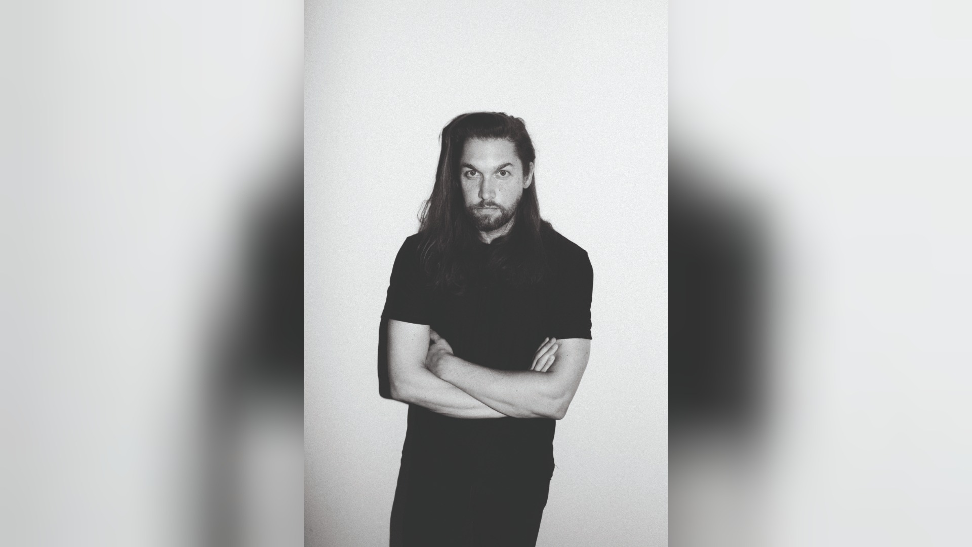 Rasmus Syman, ein Mann mit langen Haaren und Bart, steht mit verschränkten Armen vor hellem Hintergrund