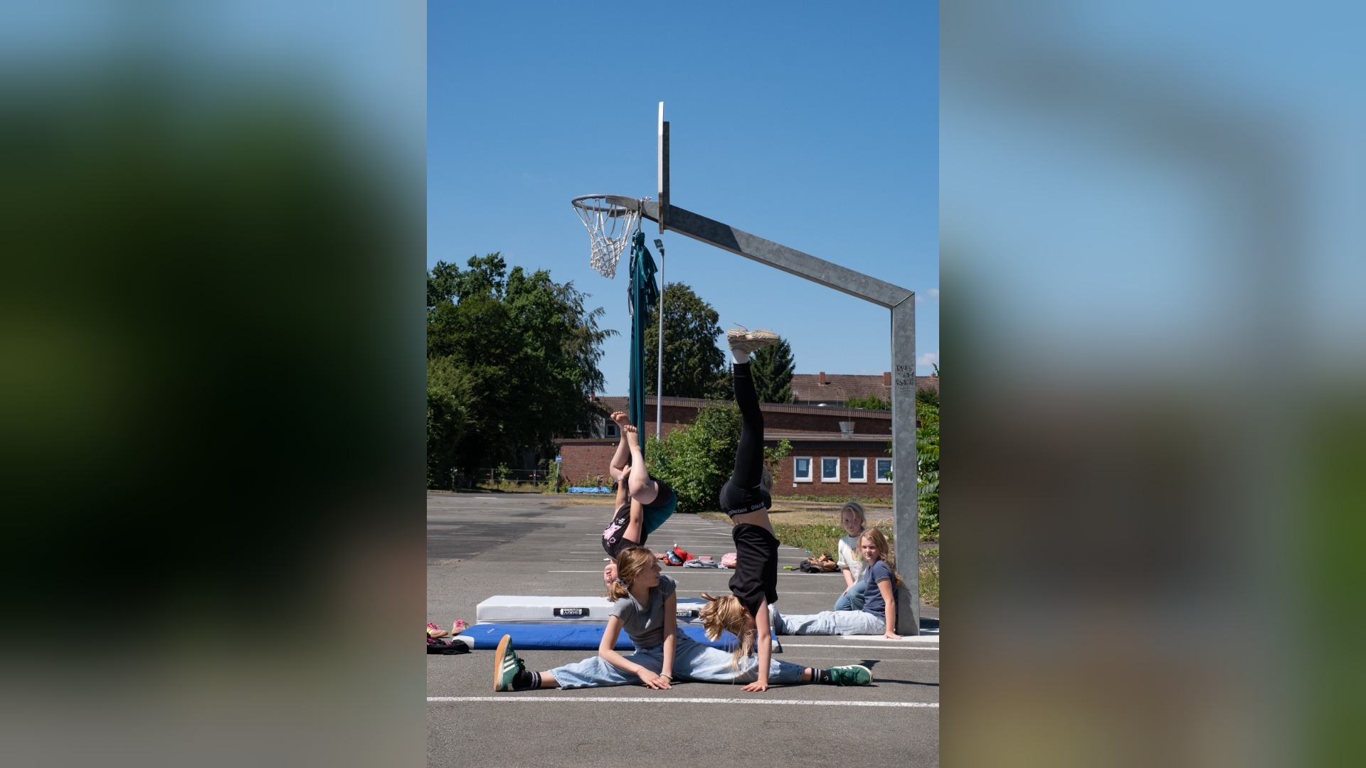 Mädchen machen Handstand, Spagat und hängen kopfüber auf einem asphaltierten Sportplatz vor einem Basketballkorb
